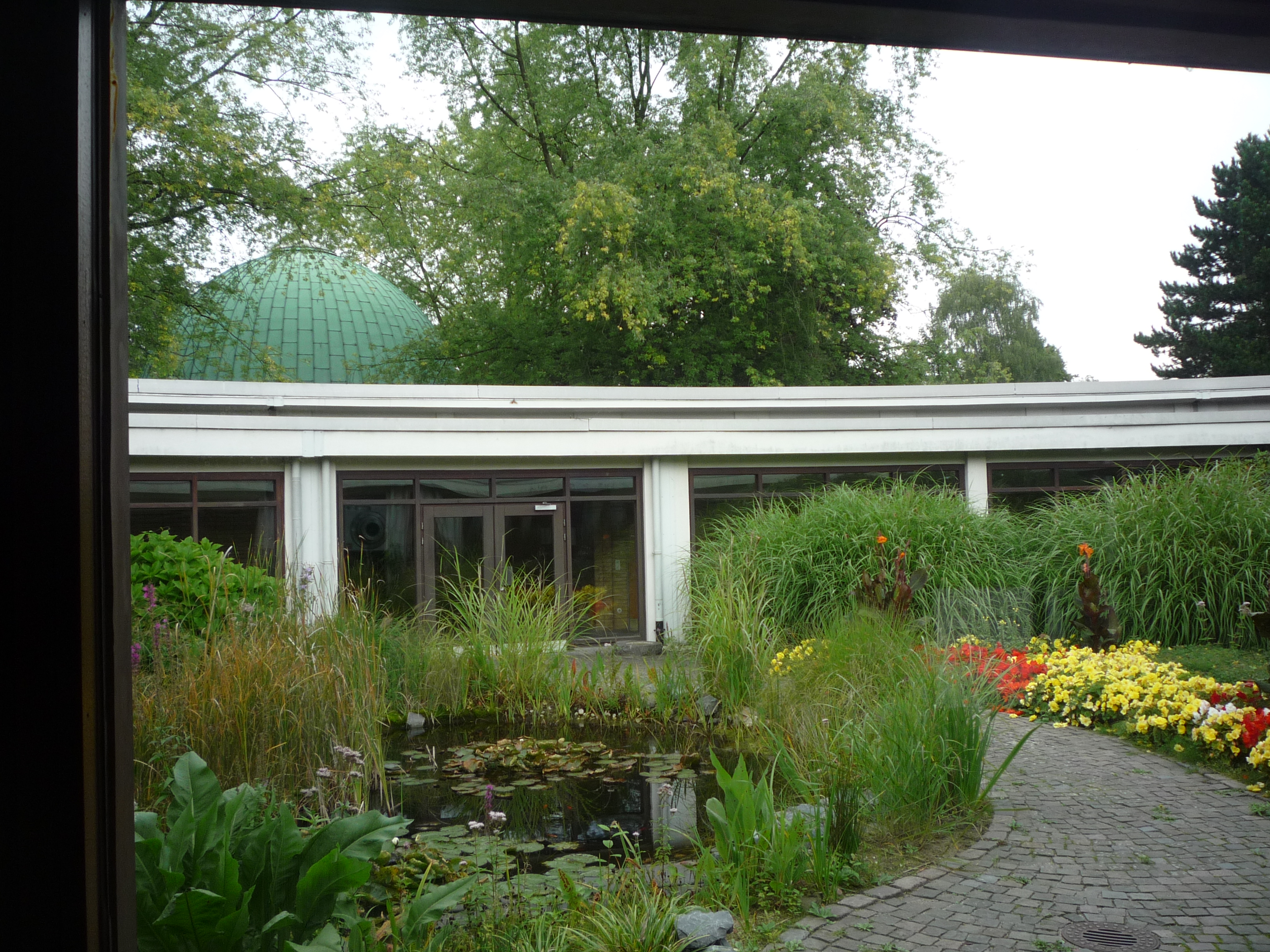 Blick in eine sonnigen runden verglasten Innenhof mit Teich, Schilf, Blumen, im Hintergrund eine kupfergrüne Kuppel.